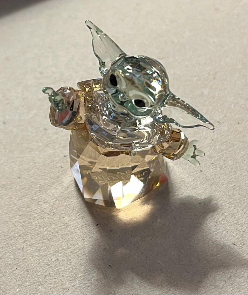 Puede incluir: Una figura de cristal transparente de un personaje con orejas y ojos grandes, sobre una base facetada. La figura tiene los brazos extendidos y est&aacute; hecha de m&uacute;ltiples piezas de cristal. La base es de color &aacute;mbar claro.