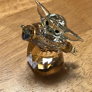 Puede incluir: Una figura de cristal de un personaje con orejas grandes y ojos grandes, en una pose con un brazo extendido. La figura est&aacute; hecha de cristal transparente y &aacute;mbar, con una base y cabeza facetadas.