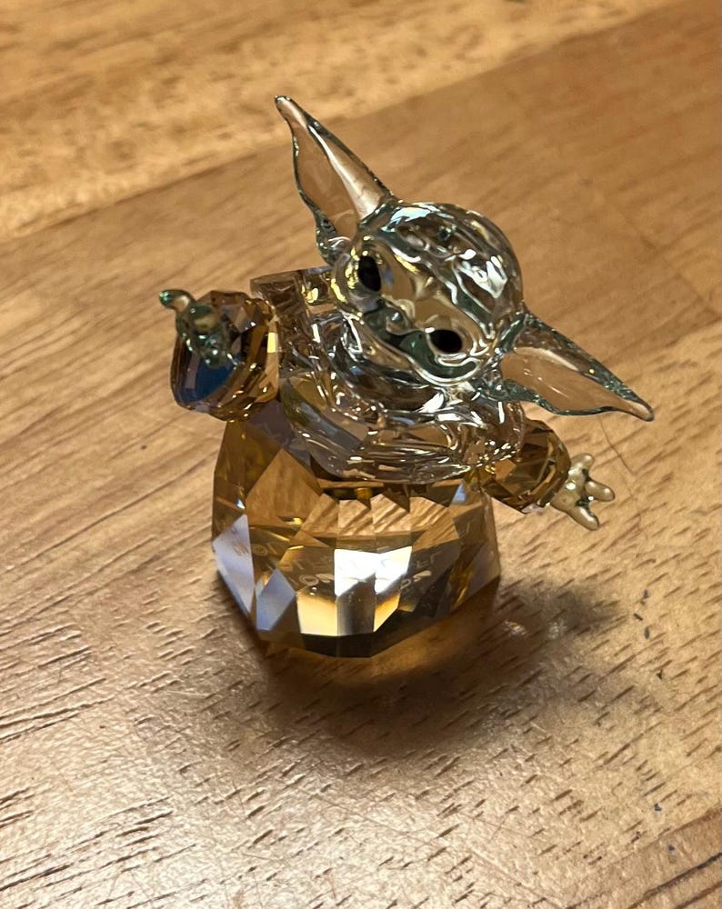 Puede incluir: Una figura de cristal de un personaje con orejas grandes y ojos grandes, en una pose con un brazo extendido. La figura est&aacute; hecha de cristal transparente y &aacute;mbar, con una base y cabeza facetadas.