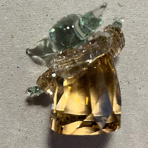 Puede incluir: Una figura de cristal facetada con forma de personaje con cabeza y orejas grandes. La cabeza y las orejas son de color verde transl&uacute;cido, mientras que el cuerpo es de color &aacute;mbar facetado. La figura est&aacute; sobre un fondo neutro.