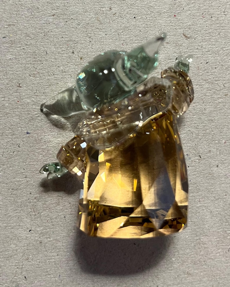 Puede incluir: Una figura de cristal facetada con forma de personaje con cabeza y orejas grandes. La cabeza y las orejas son de color verde transl&uacute;cido, mientras que el cuerpo es de color &aacute;mbar facetado. La figura est&aacute; sobre un fondo neutro.