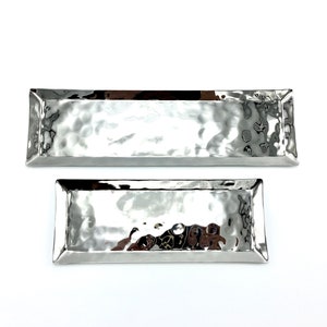 May include: Two rectangular, hammered-metal serving trays. The trays are silver-colored with a textured surface and a slightly raised edge. The trays are different sizes, with the larger tray on top and the smaller tray below.