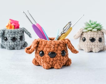 May include: Three crocheted dog-shaped containers, one brown, one gray, and one beige. The brown container is holding a set of crochet hooks and scissors.