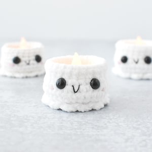 May include: Three handmade white crochet candle holders with smiling faces. Each holder has black button eyes, a stitched mouth, and a soft pink blush. The candle holders are lit with a warm, flickering light.