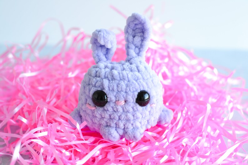 Purple chenille yarn crocheted bunny with tall ears standing in a pile of pink easter grass.