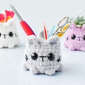 May include: Three handmade crochet cat-shaped containers in white, gray, and lavender. The gray cat holds scissors, crochet hooks, and other craft tools. The white cat holds flowers, and the lavender cat holds a small succulent. Each cat has black eyes and whiskers.