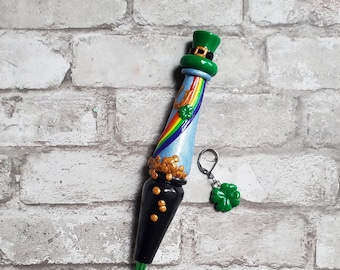 May include: A green crochet hook with a black handle decorated with a leprechaun hat, a rainbow, and gold coins. A green shamrock charm is attached to the hook.