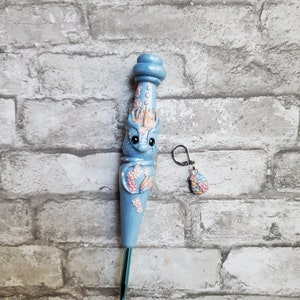 May include: A light blue crochet hook with a floral design and a matching charm. The hook has a light blue handle with a floral design and a light blue hook. The charm is a light blue flower with a light blue stem.
