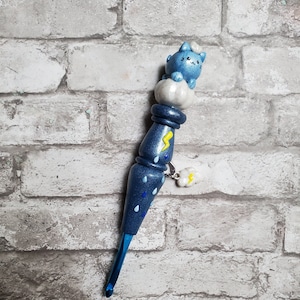May include: A blue crochet hook with a blue cat sitting on a white cloud. The hook is decorated with blue raindrops and a yellow lightning bolt.