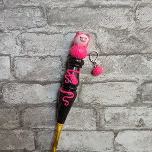 May include: A black and gold crochet hook with a pink pig and yarn ball charm. The hook has a sparkly black finish and a pink swirl design.