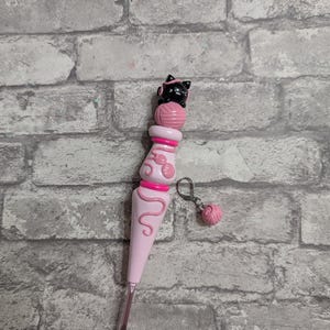 May include: A pink crochet hook with a black cat topper wearing pink glasses and a pink yarn ball. The hook has a pink handle with a decorative swirl design and a small pink yarn ball charm.