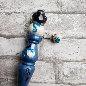 May include: A blue and white ceramic crochet hook with a black bird on top. The hook has a white bead with a blue swirl design and a small white bead with a blue swirl design hanging from it.