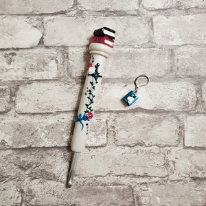 May include: A white crochet hook with a stack of miniature books on top, decorated with painted flowers and vines. A small blue book charm hangs from a silver key ring.