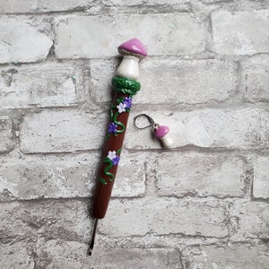 May include: A brown crochet hook with a mushroom-shaped handle. The handle is decorated with green vines, purple and white flowers, and a pink mushroom cap.
