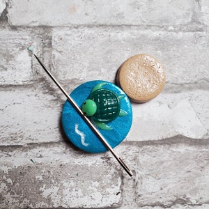 May include: A blue and green turtle needle minder with a wave design. The needle minder is on a white brick background.