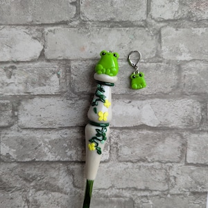 May include: A green crochet hook with a white handle decorated with green vines and yellow flowers. The top of the handle is shaped like a frog. A small green frog charm is attached to a silver hook.