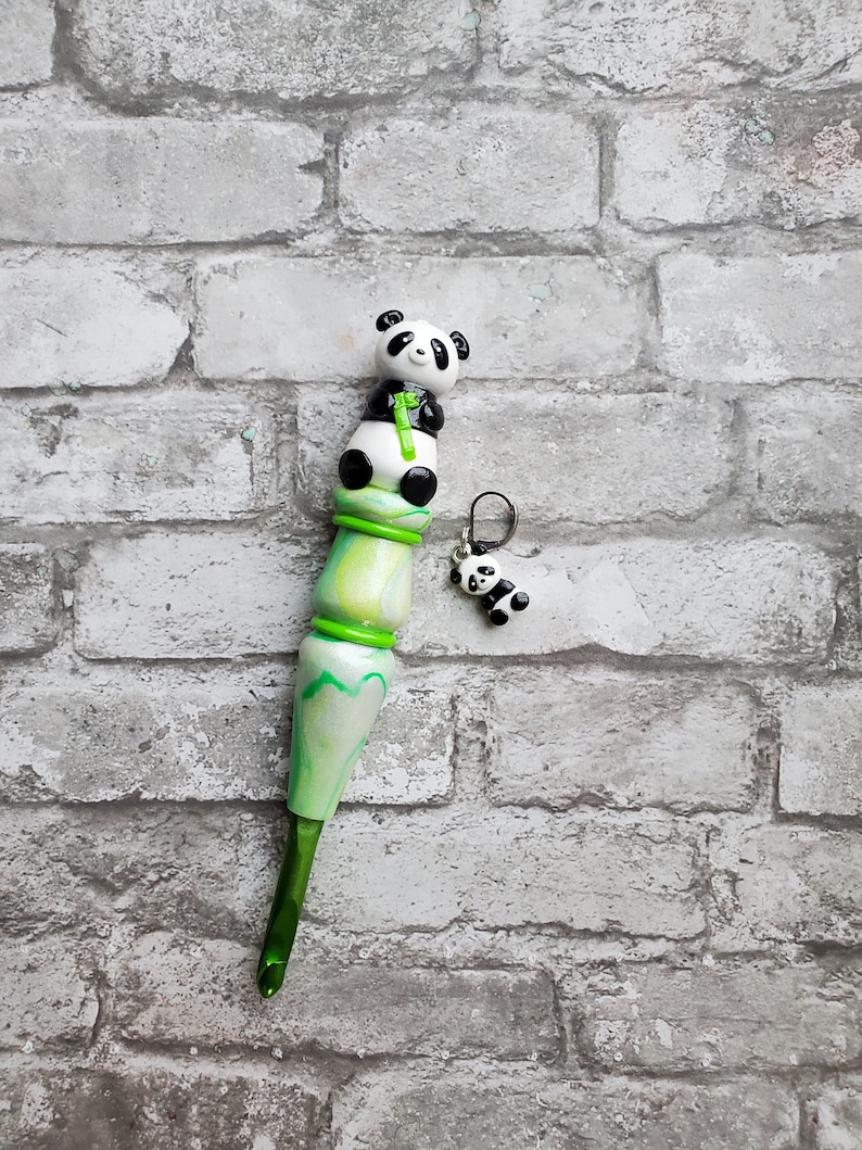 May include: Green and white crochet hook with a panda bear charm. The hook is made of wood and has a green and white swirl pattern. The panda bear charm is attached to the hook with a silver ring.