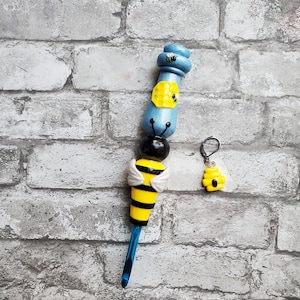 May include: A blue and yellow crochet hook with a bee design. The hook has a black and yellow bee with white wings and a honeycomb design. The hook comes with a matching bee charm.