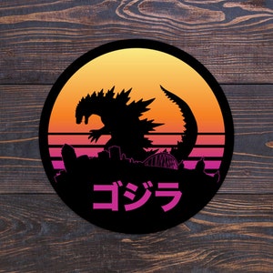 May include: A black and white silhouette of Godzilla standing in front of a city skyline with a retro sunset behind it. The text "ゴジラ" is written in pink Japanese characters below the image.