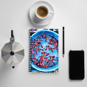 May include: A spiral bound notebook with a watercolor illustration of a blue bowl filled with red goldfish. The notebook is on a white surface with a coffee cup, a pencil, and a black smartphone.