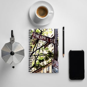 May include: A spiral bound notebook with a colorful image of a street sign that reads "Pineapple St. Henry". The sign is on a metal pole with a green tree in the background. The notebook is on a white surface with a coffee cup, a pencil, and a black smartphone.