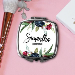 May include: A compact mirror with a floral design and the name "Samantha Bridesmaid" printed on it. The mirror is silver and has a floral design with pink, blue, and green flowers. The mirror is sitting on a pink surface.