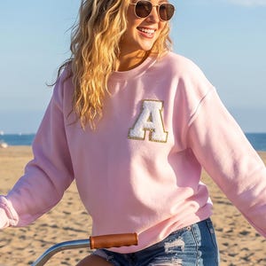 May include: A light pink long-sleeve crewneck sweatshirt featuring a large white letter A patch with a gold glitter border on the chest. The sweatshirt has a ribbed collar, cuffs, and hem, and is worn with blue denim shorts.