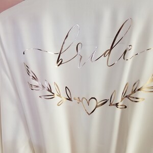 May include: Two white satin robes with gold foil lettering. One robe says "bride" with a heart and leafy branches. The other robe says "maid of honor" with a heart.