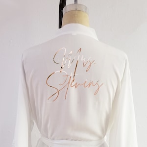 May include: White satin robe with a lace trim and the text "Mrs. Stevens" in rose gold lettering on the back.