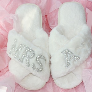 May include: White faux fur slippers with a criss-cross strap design. One slipper has "MRS" spelled out in rhinestones, the other has a single "A" in rhinestones.