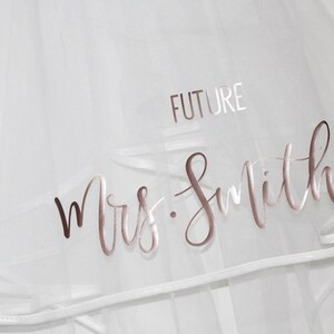 May include: White bridal veil with the words "Future Mrs. Smith" written in rose gold lettering.