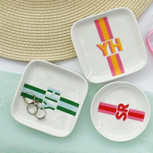 May include: Three white ceramic ring dishes with colorful stripes and initials. The dishes are on a green and white surface. The initials are "YH", "SR", and "EH".
