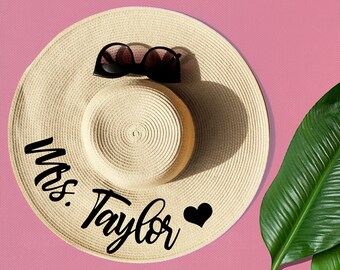 May include: A straw sun hat with a black heart and the text "Mrs. Taylor" written in black script. The hat is on a pink background with a green leaf.