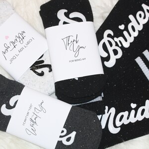 K&ouml;nnte beinhalten: Vier Paar schwarze und wei&szlig;e Socken mit wei&szlig;em Text auf den Socken. Die Socken haben eine glitzernde Textur. Der Text auf den Socken lautet "Brides", "Maid", "I can't say 'I do' without you!" und "Thank you for being my".