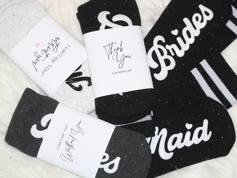 K&ouml;nnte beinhalten: Vier Paar schwarze und wei&szlig;e Socken mit wei&szlig;em Text auf den Socken. Die Socken haben eine glitzernde Textur. Der Text auf den Socken lautet "Brides", "Maid", "I can't say 'I do' without you!" und "Thank you for being my".