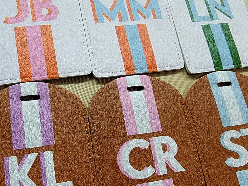 May include: A collection of rectangular and rounded luggage tags made of textured material. The top row features white tags with vertical stripes and initials JB, MM, and LN. The bottom row shows brown tags with vertical stripes and initials KL, CR, and S.