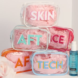 May include: Five clear plastic zippered pouches with pink tissue paper inside. Each pouch has a different colored word on the front: SKIN, AFT, FACE, AFT, TECH. Two bottles of skincare products are in the foreground.