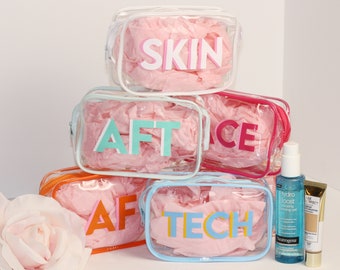 May include: Five clear plastic zippered pouches with pink tissue paper inside. Each pouch has a different colored word on the front: SKIN, AFT, FACE, AFT, TECH. Two bottles of skincare products are in the foreground.