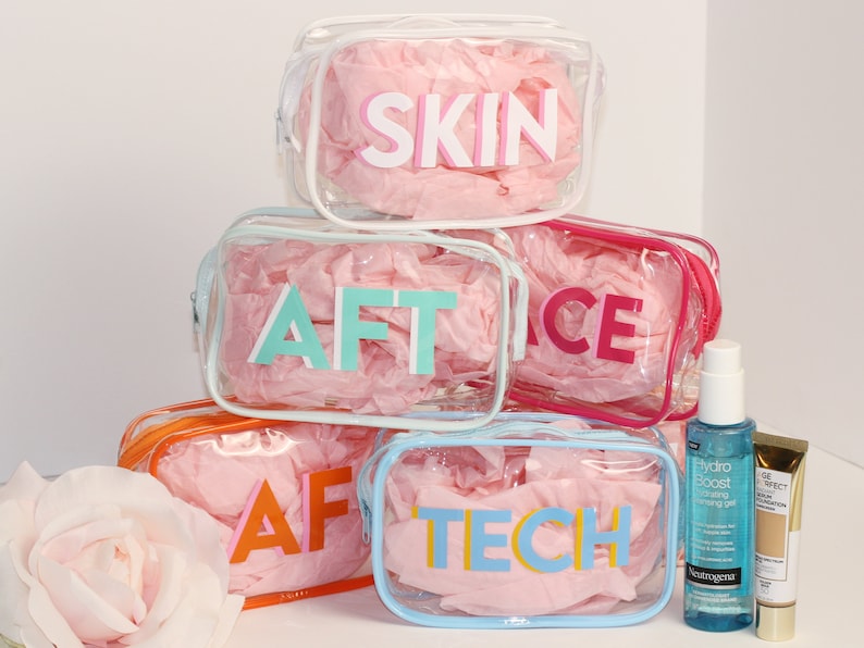 May include: Five clear plastic zippered pouches with pink tissue paper inside. Each pouch has a different colored word on the front: SKIN, AFT, FACE, AFT, TECH. Two bottles of skincare products are in the foreground.