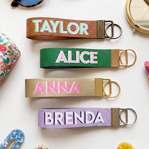 May include: Four personalised keychains in brown, green, gold, and lavender. Each keychain features a name in a contrasting colour: Taylor, Alice, Anna, and Brenda. The keychains have metal hardware and are displayed on a white surface.