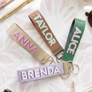 May include: Four personalised keychains in tan, brown, green, and lavender. Each keychain has a name printed in a contrasting colour: Anna, Taylor, Alice, and Brenda. The keychains have gold-coloured metal hardware.