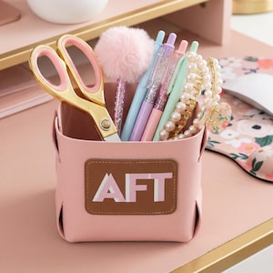 May include: A pink desk organizer with gold scissors, pens, and a fluffy pen. The organizer has a brown patch with the letters "AFT" in white. A pearl necklace and a floral hair clip are also visible.