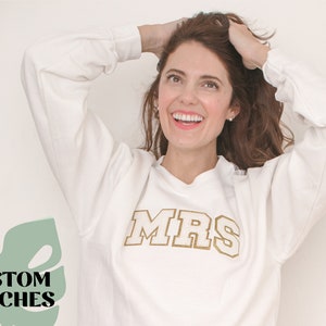 May include: A white sweatshirt with a gold "MRS" patch on the front. The sweatshirt is worn by a person with brown hair who is smiling and holding their hair up.