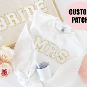 May include: White sweatshirt with a gold glitter "MRS" patch. The sweatshirt is laying on a white surface with a tote bag that says "BRIDE" in gold glitter and some pink flowers.