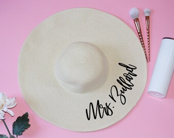 May include: A large, beige sun hat with a wide brim and a black inscription that reads "Mrs. Ballard".