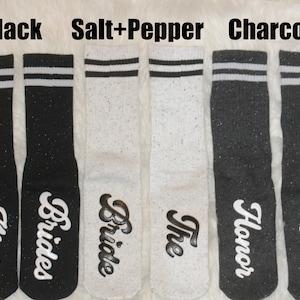 K&ouml;nnte beinhalten: Sechs Paar Socken in Schwarz, Wei&szlig; und Grau mit wei&szlig;en Streifen. Die Socken haben unterschiedliche Texte, darunter "Bride", "Bridesmaid", "Maid of Honor" und "The Bride".