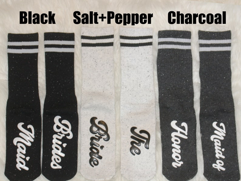 K&ouml;nnte beinhalten: Sechs Paar Socken in Schwarz, Wei&szlig; und Grau mit wei&szlig;en Streifen. Die Socken haben unterschiedliche Texte, darunter "Bride", "Bridesmaid", "Maid of Honor" und "The Bride".