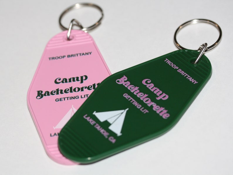 May include: Two pink and green keychains shaped like motel keys. The keychains have the text "Camp Bachelorette Getting Lit" and "TROOP BRITTANY" on them. The keychains also have a white tent graphic on them.
