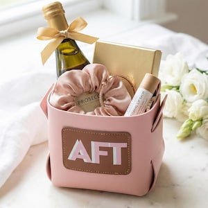 May include: A pink leatherette gift basket with the letters "AFT" on the front. The basket contains a bottle of sparkling wine with a gold bow, a gold box, a pink scrunchie, a bracelet, and a lip balm.