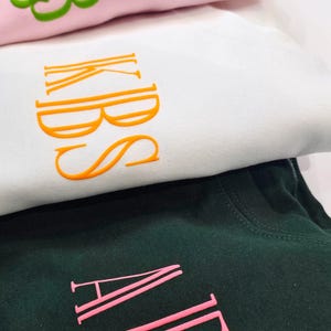 May include: Three folded t-shirts with embroidered names. The top shirt is pink with green embroidery that says "SJB". The middle shirt is white with orange embroidery that says "KIBS". The bottom shirt is dark green with pink embroidery that says "ALPI".